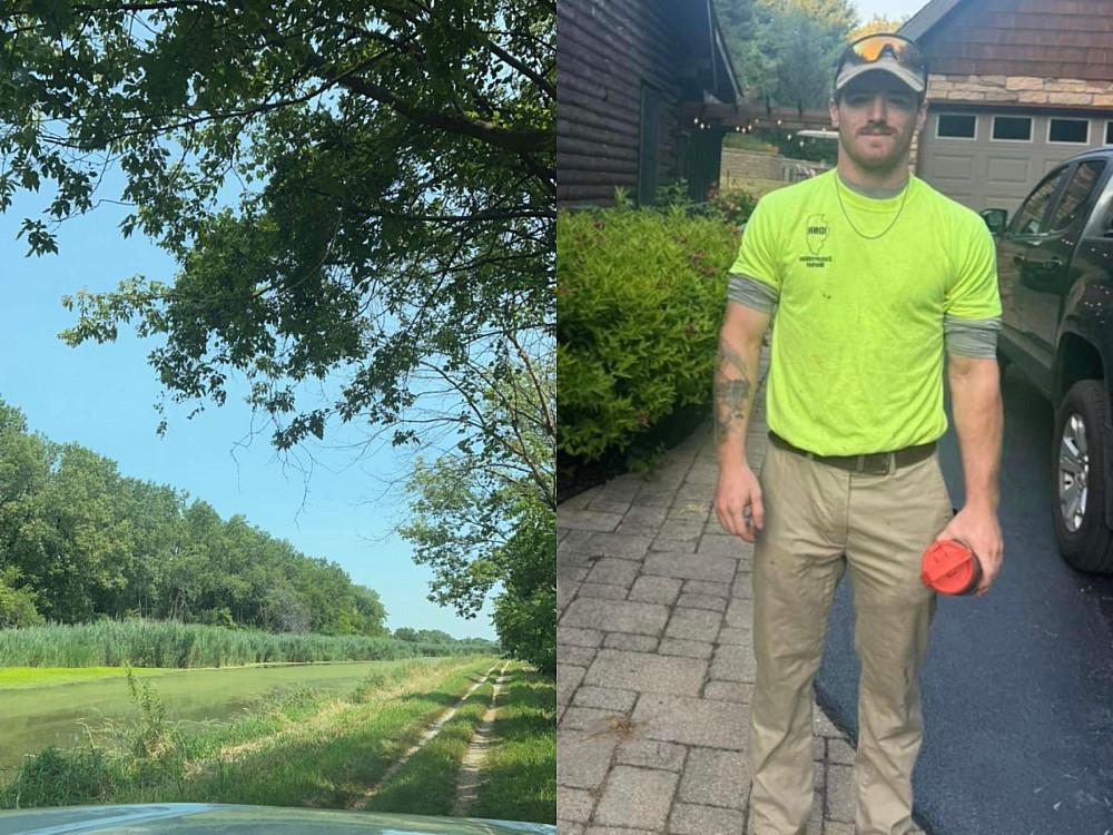 Person poses for picture in work clothes. Car driving next to trail, near trees and grass field.